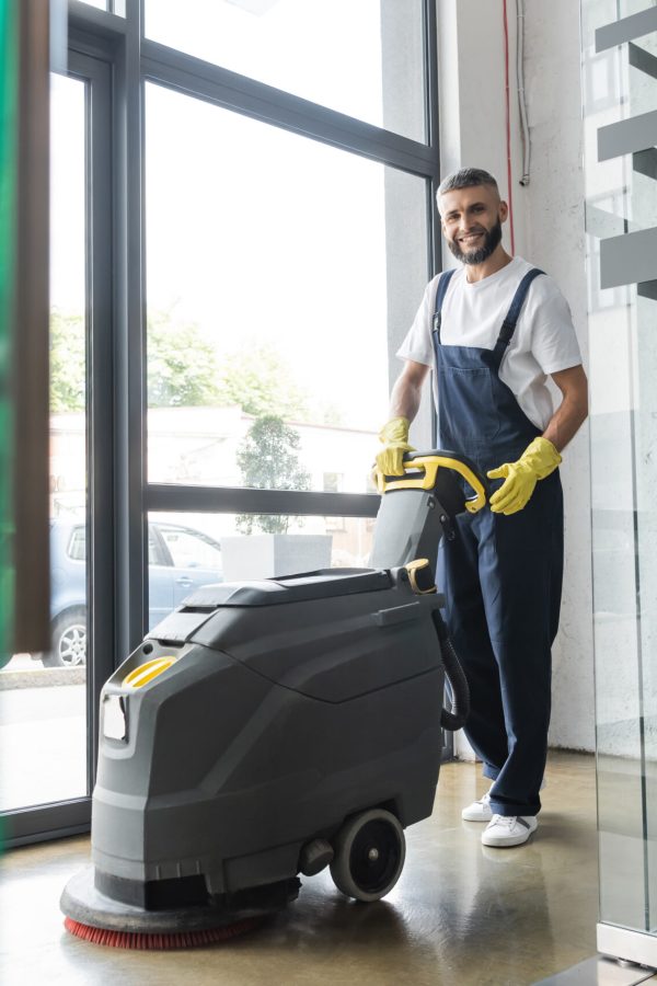 full length of professional cleaner with electrical floor scrubber machine smiling at camera